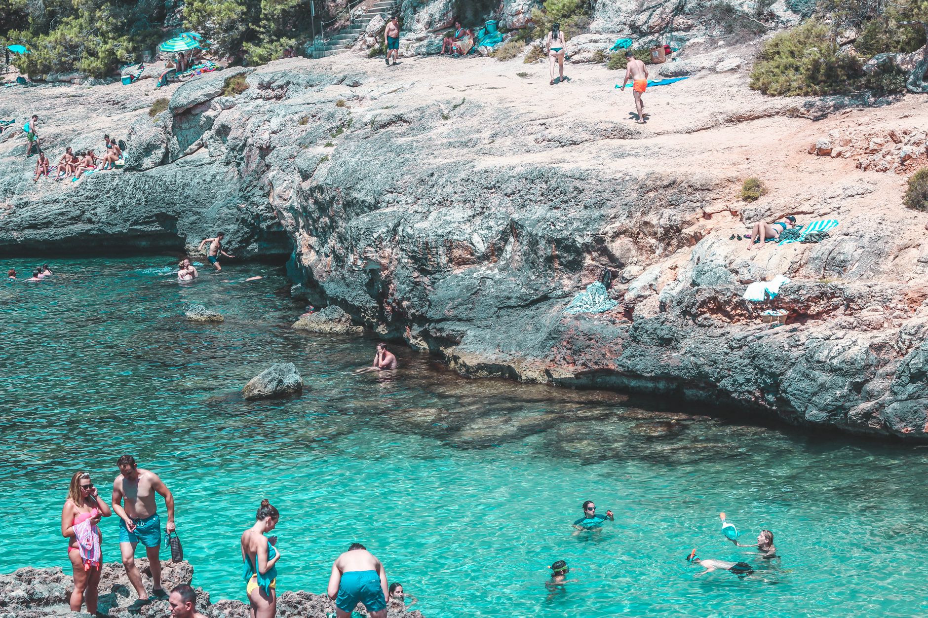 A lively scene of people swimming and relaxing by a rocky seashore under the summer sun.